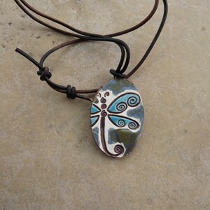 May include: An oval-shaped pendant featuring a dragonfly design with turquoise and white wings on a mottled blue and green background. The pendant is suspended from a dark brown leather cord with adjustable sliding knots for a secure fit.
