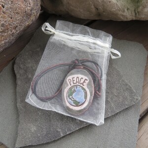May include: A hand-painted stone pendant with the word "PEACE" and a globe design, displayed in a sheer white drawstring bag. The pendant has a brown cord and is set against a gray stone surface.