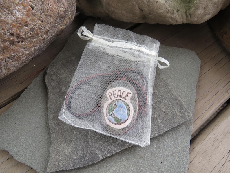 May include: A hand-painted stone pendant with the word "PEACE" and a globe design, displayed in a sheer white drawstring bag. The pendant has a brown cord and is set against a gray stone surface.