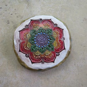 May include: A round, hand-painted stone featuring a colorful mandala design. The mandala has a central blue and purple flower surrounded by layers of red, orange, yellow, and green petals, all set against a white background.