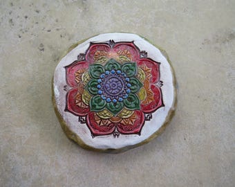 May include: A round, hand-painted stone featuring a colorful mandala design. The mandala has a central blue and purple flower surrounded by layers of red, orange, yellow, and green petals, all set against a white background.