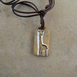May include: A handcrafted pendant necklace featuring a rectangular ceramic pendant with a giraffe design. The giraffe is depicted in white and yellow against a tan background. The necklace has a brown leather cord.