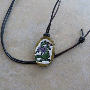 May include: A black leather cord necklace with a green and purple dragon pendant. The pendant is oval shaped and has a white background.