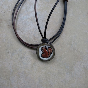 May include: A round pendant necklace featuring a brown squirrel design. The pendant is set on a light-colored background with a dark brown leather cord. The squirrel is depicted in shades of brown and white.