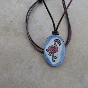 May include: An oval-shaped pendant featuring a hand-painted pink flamingo on a light blue and white background. The pendant is suspended from a dark brown and black leather cord necklace that loops through a small leather knot at the top.