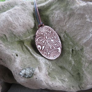 May include: Oval pendant with a brown and white intricate design, suspended by a brown leather cord. The pendant is set against a textured, gray stone background, showcasing the handcrafted details.