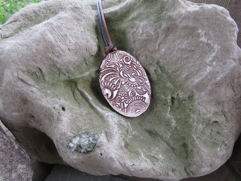 May include: Oval pendant with a brown and white intricate design, suspended by a brown leather cord. The pendant is set against a textured, gray stone background, showcasing the handcrafted details.