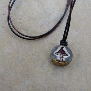 May include: A round pendant necklace featuring a mushroom design. The pendant has a gray background with a purple mushroom and a yellow base. The necklace has a black and brown leather cord.