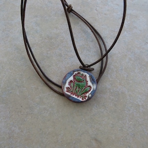 May include: A brown leather cord necklace with a round ceramic pendant featuring a green frog with red and white details.