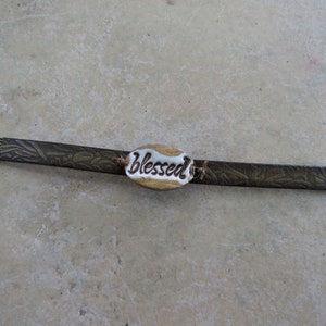 May include: A brown leather bracelet with a gold-toned metal clasp and a white ceramic charm that says "blessed".