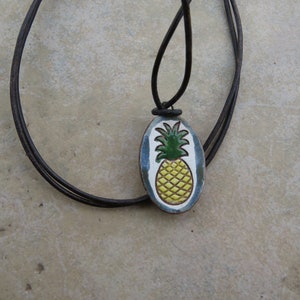 May include: A pineapple pendant necklace with a brown leather cord. The pendant is oval shaped and features a yellow pineapple with green leaves on a white background with blue accents.