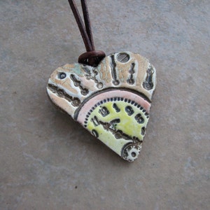May include: Heart-shaped pendant with a steampunk design. The ceramic pendant features a clock face and gears in yellow, pink, and beige tones. It hangs from a brown leather cord.