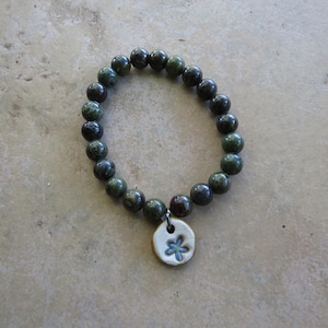 May include: A beaded bracelet featuring dark green and black round beads. A small, round, off-white charm with a blue flower design hangs from the bracelet. The bracelet is displayed on a light brown surface.