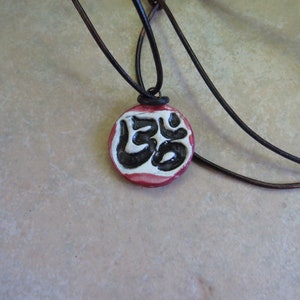 May include: A red, white, and black ceramic pendant with the symbol "Om" on it. The pendant is on a brown leather cord.