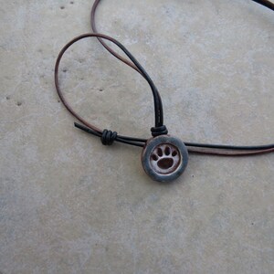 May include: A round, dark-toned ceramic pendant featuring an embossed paw print design. The pendant is suspended from a thin, adjustable cord made of two strands, one black and one brown, secured with a simple sliding knot mechanism.