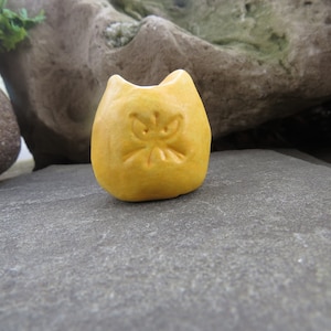 May include: A small, yellow ceramic cat figurine with a grumpy expression. The cat is sitting on a gray stone surface.