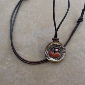 May include: A round pendant necklace with a detailed eye design. The pendant features a brown, orange, green, and white color scheme. The necklace has a black leather cord with a brown leather accent.