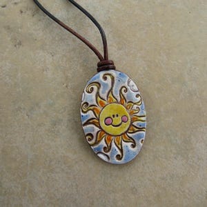 May include: Oval ceramic pendant with a smiling sun design. The sun has a yellow face with pink cheeks and orange rays, set against a blue and white background. The pendant hangs from a brown leather cord.