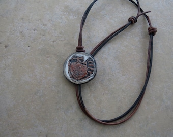 May include: A necklace featuring a round, off-white pendant with a hand-painted brown and black raccoon illustration. The pendant is suspended from a double-strand cord made of one thin black leather strip and one thin brown leather strip.
