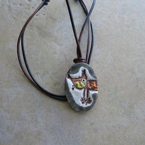 May include: Oval pendant necklace with a hand-painted design of a cat-like creature in orange, yellow, and white on a gray stone. The pendant hangs from a brown and black leather cord.