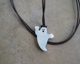 May include: A white ceramic ghost pendant with black eyes hangs from a brown leather cord. The ghost has a simple, whimsical design and is a fun and spooky accessory.