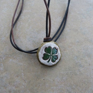 May include: A green four-leaf clover pendant on a brown and black leather cord necklace. The pendant is made of ceramic and has a white background.
