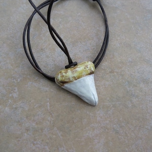May include: A white and yellow ceramic shark tooth pendant on a brown leather cord necklace.