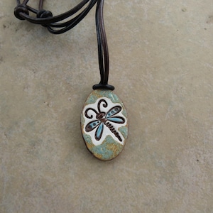 May include: An oval-shaped pendant featuring a stylized dragonfly illustration in white, light blue, and dark brown, suspended from a dark brown leather cord. The pendant has a textured, mottled surface with a mix of green, gold, and white tones.