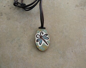 May include: An oval-shaped pendant featuring a stylized dragonfly illustration in white, light blue, and dark brown, suspended from a dark brown leather cord. The pendant has a textured, mottled surface with a mix of green, gold, and white tones.