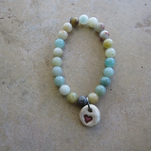 May include: A beaded bracelet with a variety of pastel-colored beads, including light blue, yellow, and white. A small, round, off-white charm with a red heart is attached. The bracelet is displayed on a neutral-toned surface.