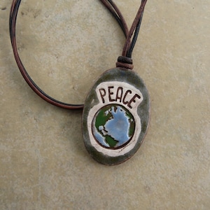 May include: A handcrafted pendant necklace featuring a ceramic oval pendant with the word "PEACE" and a globe design. The pendant is olive green with a white border and a brown leather cord.