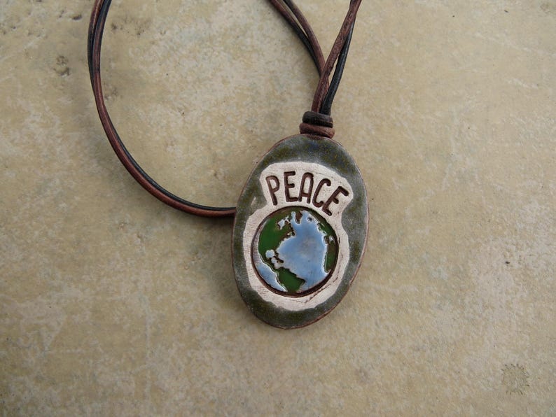 May include: A handcrafted pendant necklace featuring a ceramic oval pendant with the word "PEACE" and a globe design. The pendant is olive green with a white border and a brown leather cord.