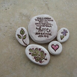May include: A collection of white, painted stones with inspirational messages and floral designs. The largest stone has the text "FOR I KNOW THE PLANS I HAVE FOR YOU, SAYS THE LORD... TO GIVE YOU A hope AND A FUTURE JER 29:11". Other stones feature a leaf, a flower, and a heart.