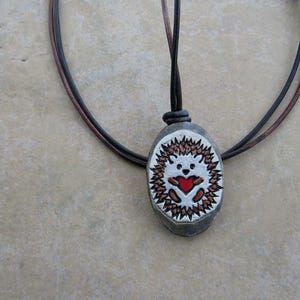 May include: A pendant necklace featuring an oval-shaped stone with a hand-painted hedgehog design. The hedgehog has a red heart, brown spikes, and a white face. The necklace has a black and brown cord.