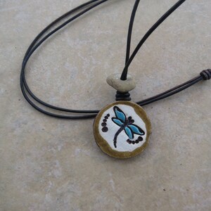 May include: A black leather cord necklace with a round ceramic pendant featuring a blue dragonfly design on a white background. The pendant is surrounded by a brown border.