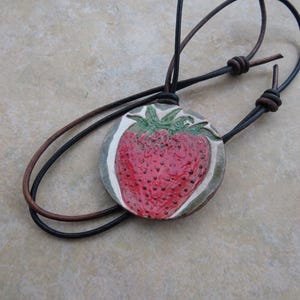 May include: A brown leather cord necklace with a round ceramic pendant featuring a hand-painted strawberry design. The strawberry is red with green leaves and a white background.