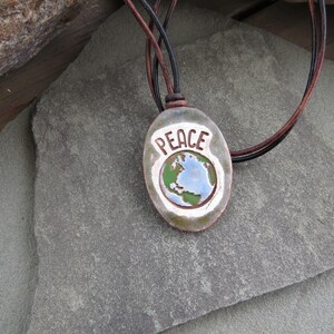 May include: A ceramic pendant necklace with the word "PEACE" in white lettering. The pendant features a globe design with blue and green colors, and is strung on a leather cord.