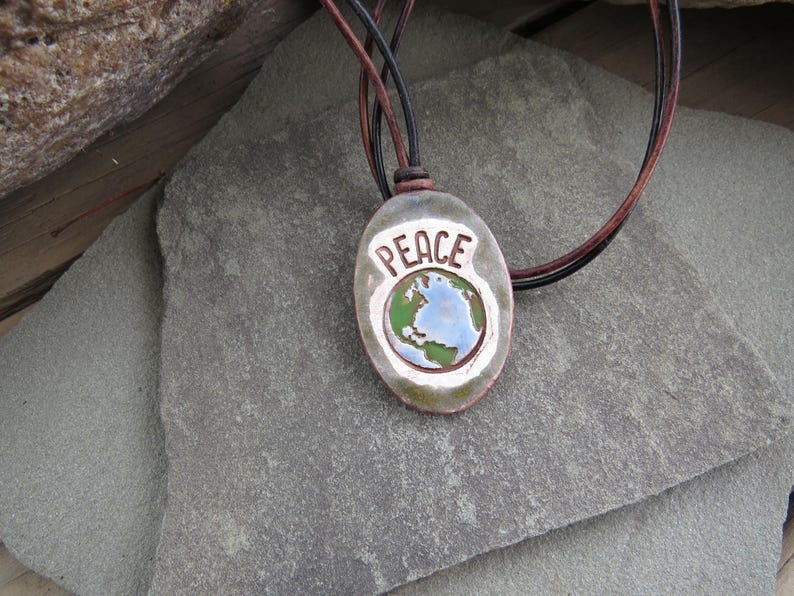 May include: A ceramic pendant necklace with the word "PEACE" in white lettering. The pendant features a globe design with blue and green colors, and is strung on a leather cord.