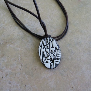 May include: A white ceramic pendant with a black and white illustration of cats, hanging from a brown leather cord.