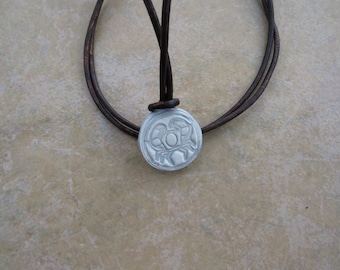 May include: A silver pendant necklace with a leather cord. The pendant features a raised design of a stylized eye with a circle in the center.