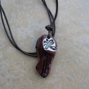 May include: A brown leather cord necklace with a red and black ceramic pendant featuring a white floral design.