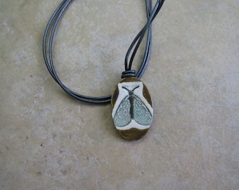 May include: A brown ceramic pendant with a white and blue moth design hangs from a black leather cord.