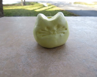 May include: A small, yellow ceramic cat figurine with a simple, carved face.