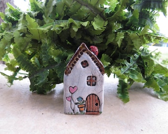 May include: A small, handmade ceramic house with a white exterior, brown roof, and a red chimney. The house features a wooden door, a small window, and pink heart decorations. A potted plant and green foliage are also visible.