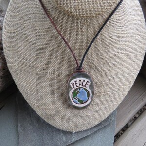 May include: A handcrafted pendant necklace featuring a stone pendant with the word "PEACE" and a globe design. The pendant hangs from a leather cord. The necklace is displayed on a burlap-covered bust.