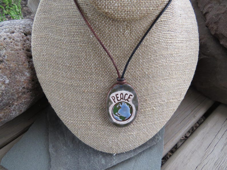 May include: A handcrafted pendant necklace featuring a stone pendant with the word "PEACE" and a globe design. The pendant hangs from a leather cord. The necklace is displayed on a burlap-covered bust.