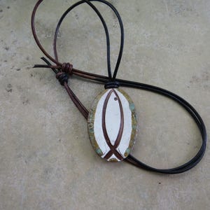 May include: An oval-shaped pendant featuring a white center with a brown fish-like graphic, surrounded by a textured border with green and gold tones. The pendant hangs from a dark brown leather cord with an adjustable sliding knot closure.