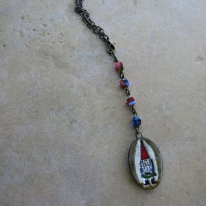 May include: A whimsical necklace featuring a pendant with a hand-painted gnome design. The oval pendant hangs from a dark chain, accented with colorful beads. The gnome wears a red hat and a blue outfit.