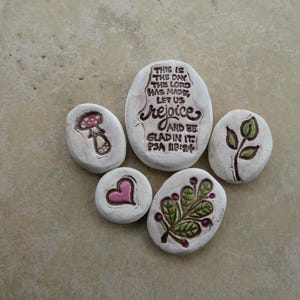 May include: Five white painted stones with various designs. One stone has a mushroom, another a pink heart, and a third a sprig of leaves. The largest stone has the text "THIS IS THE DAY THE LORD HAS MADE, LET US REJOICE AND BE GLAD IN IT! PSA 118:24".