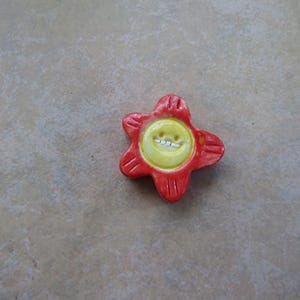 May include: A red ceramic flower with a yellow center. The center has a smiley face design.